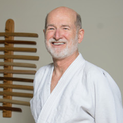 Headshot of Dave Becker, a white man in his 60s with short gray hair and a white beard, smiling and wearing a white gi jacket.