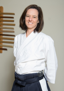 Headshot of Malory Graham, a white woman in her 50s with shoulder-length brown hair, smiling and wearing a white gi jacket and a blue hakama. 