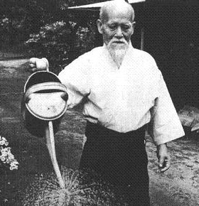 The image shows Morehei Ueshiba, or O Sensei, the founder of Aikido, an older Japanese man with a white beard and mustache, wearing a white gi and hakama, holding a large watering can and pouring water on plants. 
