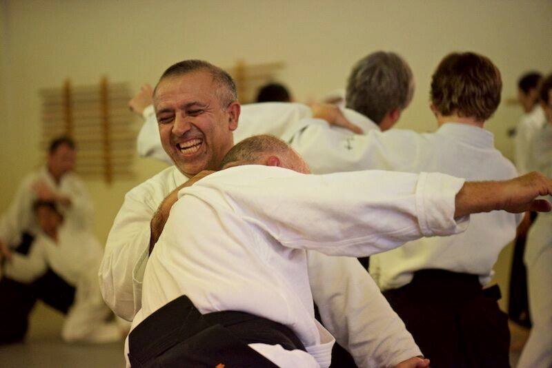 The image shows two middle-aged men at an aikido seminar. A man with graying hair is smiling and laughing as he throws another man with graying hair. There are pairs of people throwing each other in the background.