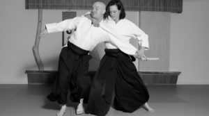 Malory Graham taking away a tanto (wooden practice knife) from Pat Roux. She is behind him with one hand around his neck holding on to his gi collar and the other hand holding the hand with the tanto. They are wearing white gis and blue hakama. 