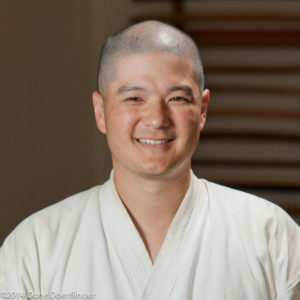 Headshot of John Peng, an Asian man in his 30s with a shaved head, smiling and wearing a white gi jacket.