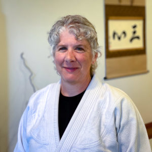 Joanne Veneziano, a white woman wearing glasses with curly gray hair, wearing a white gi standing in front of calligraphy.
