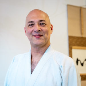 A smiling white man with a shaved head standing in front of calligraphy wearing a white gi.
