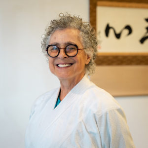 Joanne Veneziano, a smiling white woman wearing glasses with curly gray hair, wearing a white gi standing in front of calligraphy.