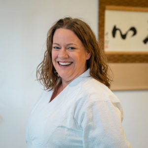A smiling white woman with shoulder length light brown hear standing in front of calligraphy wearing a white gi.