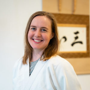 A smiling white woman with light brown hair standing in front of calligraphy wearing a white gi.